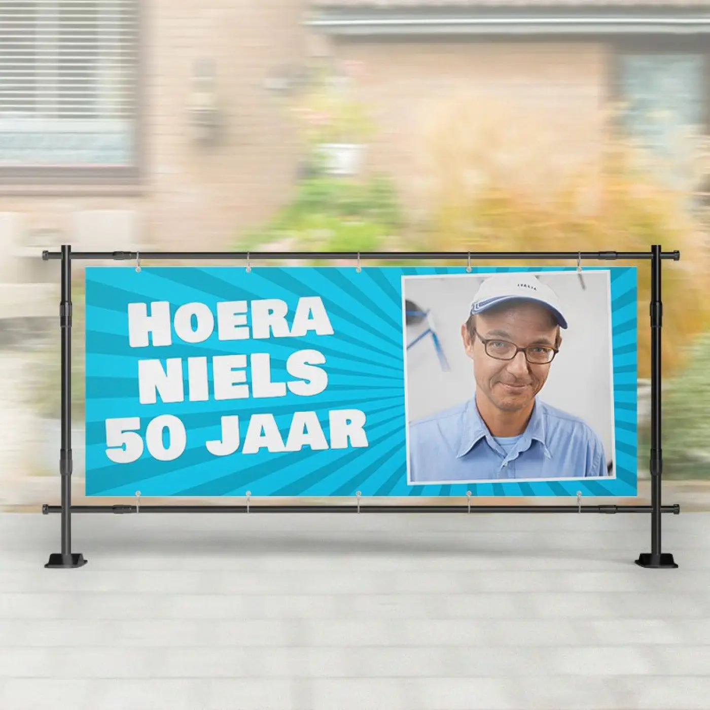 Blauw spandoek abraham verjaardag met witte tekst hoera niels 50 jaar en foto van man op zwart metalen frame